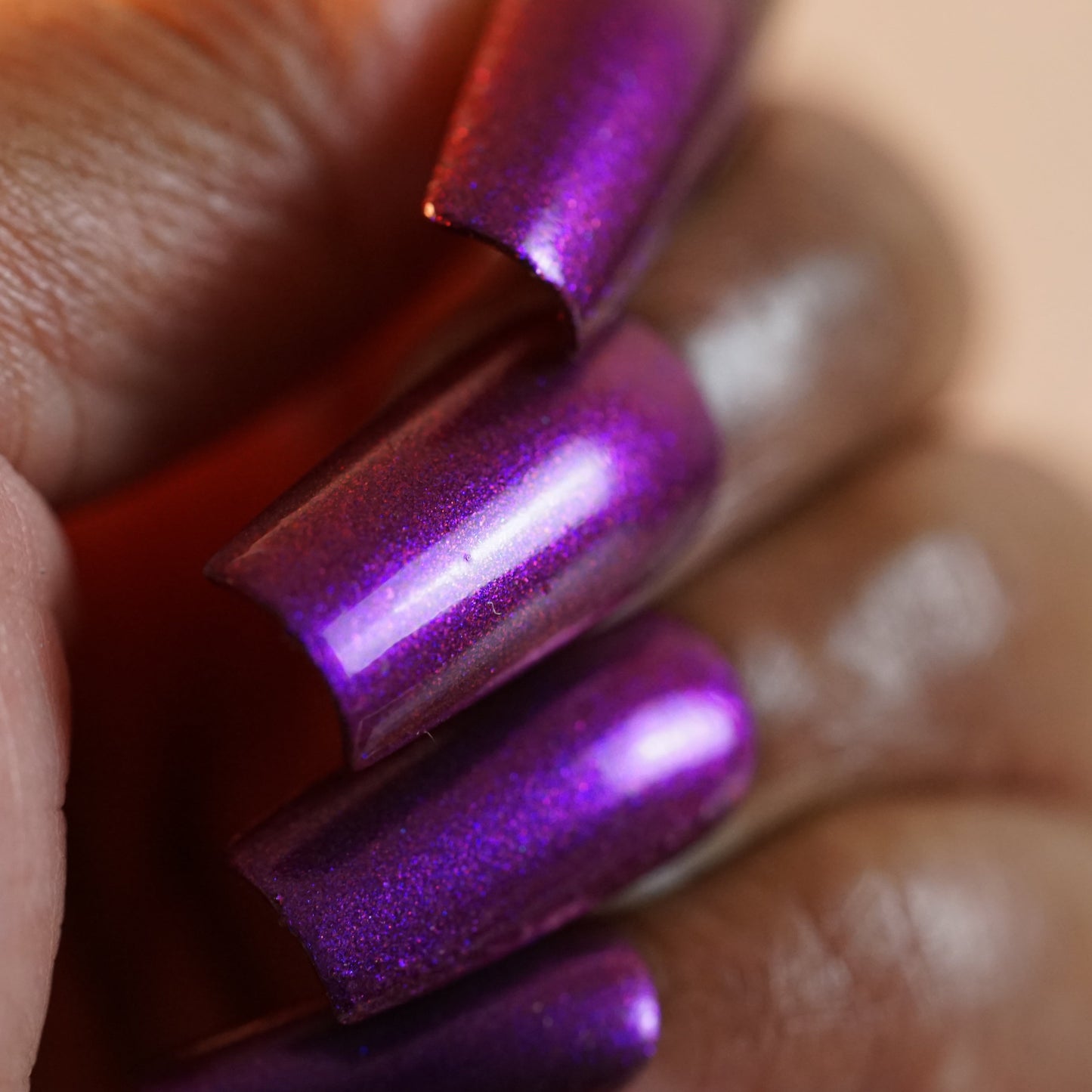 A hand displays long, shimmering purple nails with a metallic finish and pointed tips gleaming under the light. Achieve this look with PI Colors' "Danbury Does It.000," a purple multichrome nail polish offering a 10-free formula.