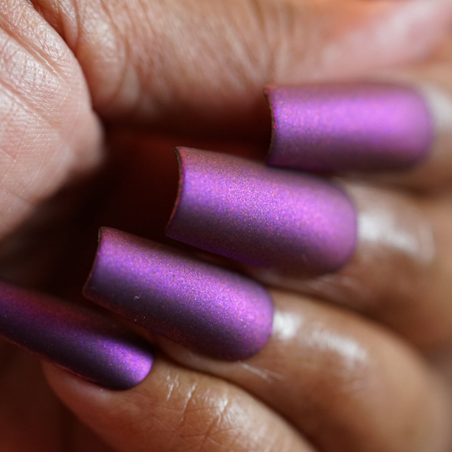 Close-up of a hand with long, matte nails featuring a metallic sheen from PI Colors' Danbury Does It.000. The multichrome polish shifts from purple to blue on neatly manicured nails, enhancing the medium brown skin tone.