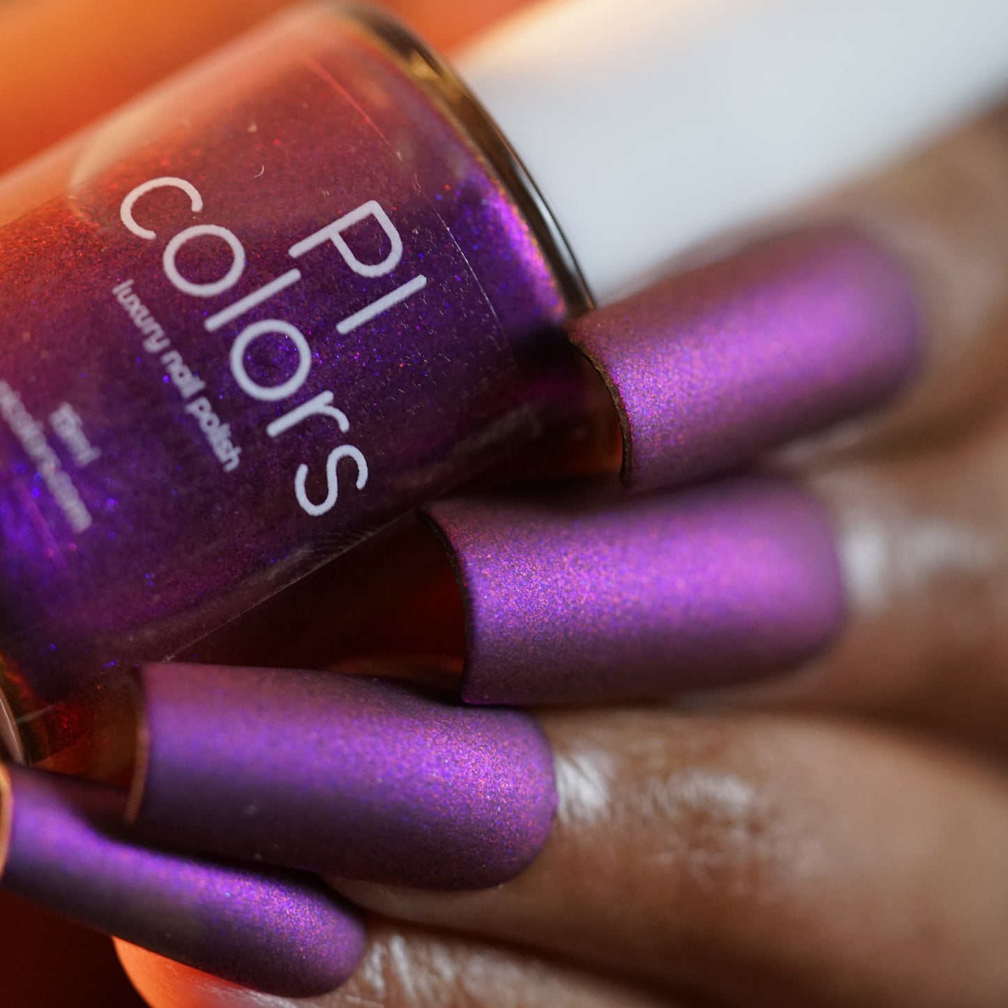 A close-up features a hand with long nails painted in PI Colors' Danbury Does It.000, a stunning purple multichrome polish. The gleaming texture is highlighted by the lighting, emphasizing the rich allure of this luxurious 10-free formula.