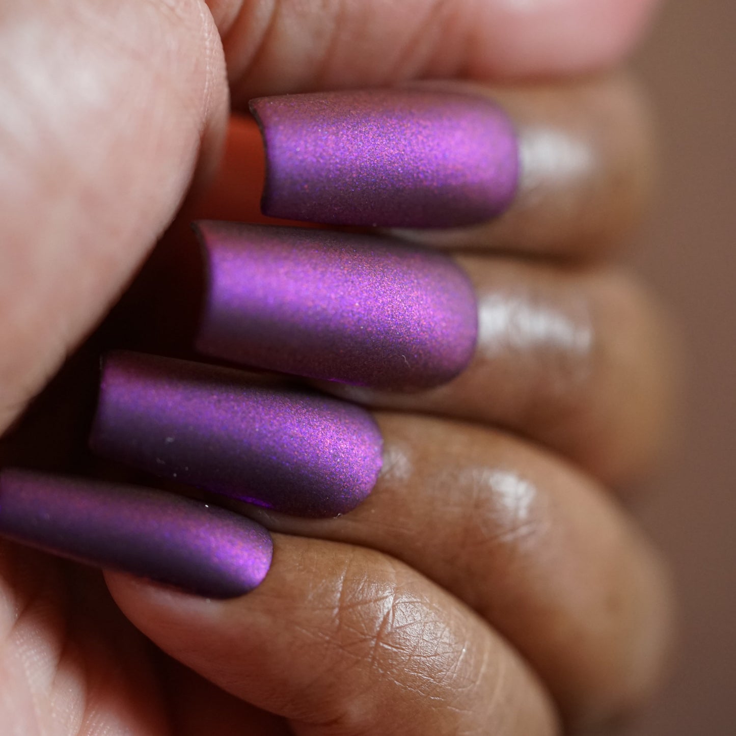 A close-up shows a hand flaunting long, neatly manicured nails painted in metallic purple multichrome with "Danbury Does It.000" by PI Colors. The medium-dark skin tone enhances the smooth, rounded nails featuring a sleek matte finish.