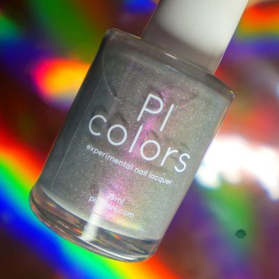 A bottle of PI Colors Cirrus.118, a 10-free nail lacquer, features a pink gray polish with a purple shimmer set against a vibrant backdrop that reflects rainbow-like colors.
