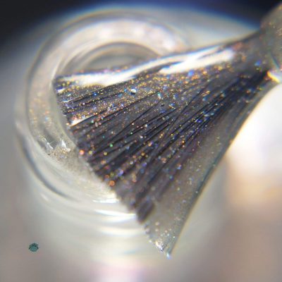 A close-up view of a nail polish brush reveals the sparkling, glittery silver hue of PI Colors' Cirrus.118 Pink Gray Nail Polish. The brush is suspended over the open mouth of a small bottle, coated in the brand's shimmering liquid 10-free formula, set against a softly blurred background.