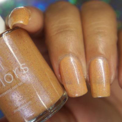 A close-up of a hand with long nails painted in PI Colors' Sunrise Tellin.027 Pale Orange Nail Polish shows off the shade's shimmery peachy-orange essence. The polish bottle is held beside the nails, reflecting the same captivating hue. A softly blurred multicolored background adds a dreamy touch to the scene.