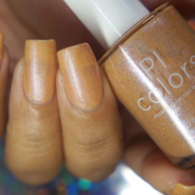 A hand with long nails is adorned in a shimmery pale orange polish called "Sunrise Tellin," accentuated by its holographic glitter. The nail polish bottle, labeled "PI Colors," is elegantly held in the same hand against a blurred, multicolored background.