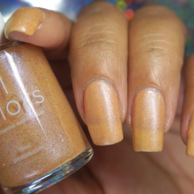 A close-up shot captures a hand adorned with long nails coated in Sunrise Tellin.027, a shimmery pale orange polish by PI Colors, featuring subtle holographic glitter. The bottle is nestled snugly between the fingers, displaying both the label and coordinating shade. A blurred, colorful backdrop enhances the vibrant appearance of the scene.
