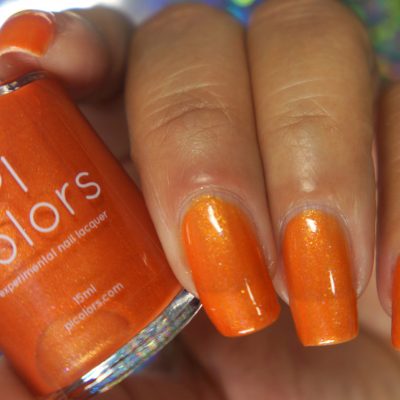 A hand with orange-painted nails holds a bottle of "Sun Conure.097 Orange Nail Polish" from PI Colors. The glossy, well-manicured nails capture the vibrant hue of this luxury 10-free polish perfectly.