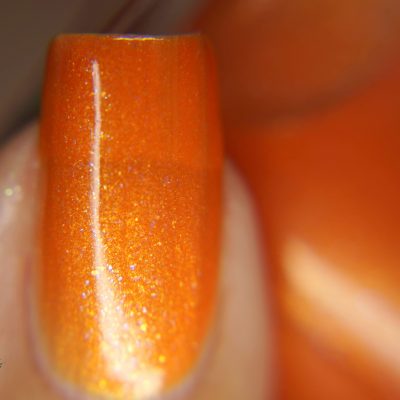 A close-up of a fingernail adorned with PI Colors' Sun Conure.097 Orange Nail Polish showcases its vibrant orange hue, complete with a shimmery, metallic finish. This 10-free polish adds a touch of luxury, while the blurred background emphasizes the glossy texture and sparkling effect of this stunning nail color.