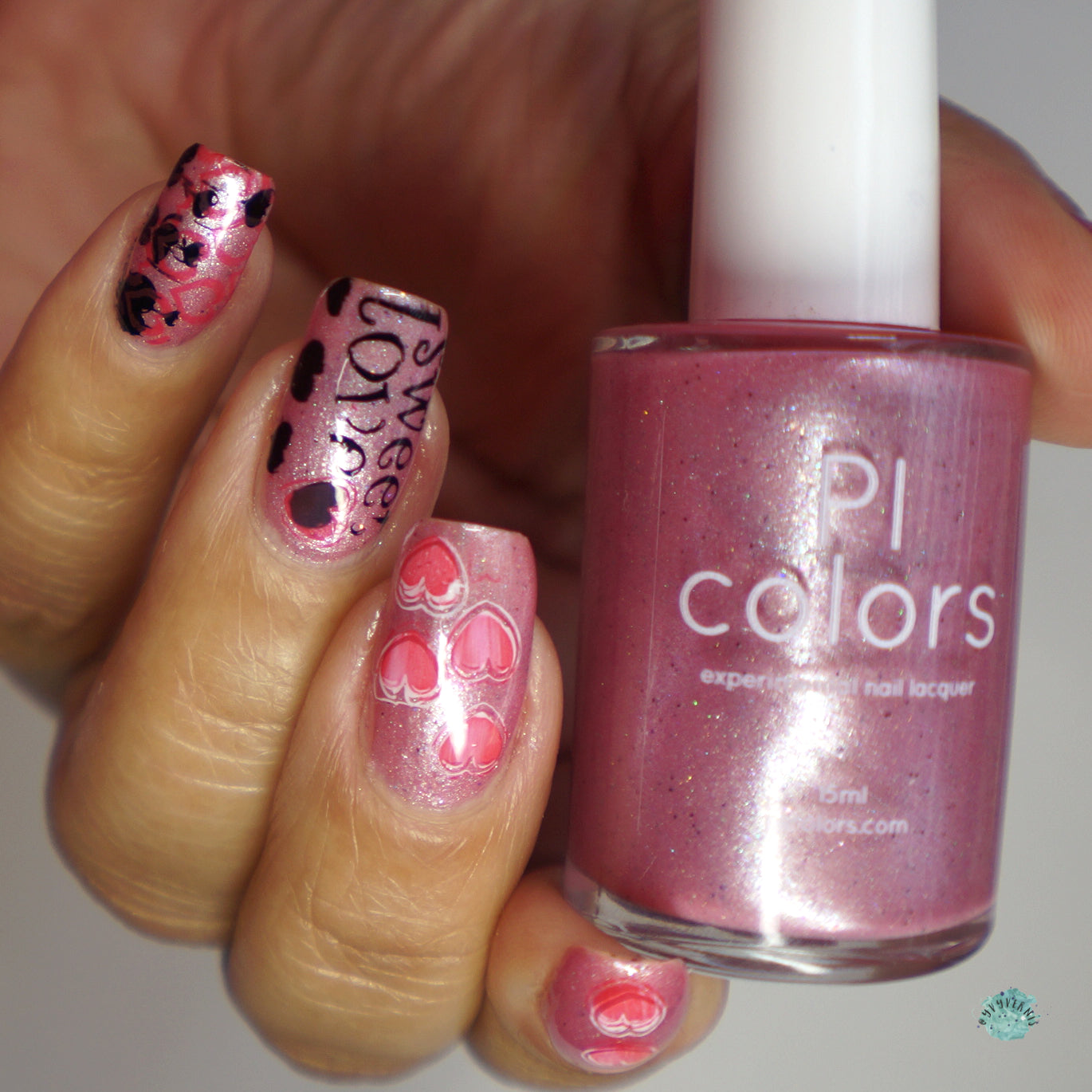 Close-up of a hand with shimmering pink nail polish and heart designs on the nails. One nail features the text "love" with black hearts, enhanced by holographic glitters. The person holds a bottle of Roseate Bliss.012 Pink Nail Polish by PI Colors, which is 10-free lacquer.