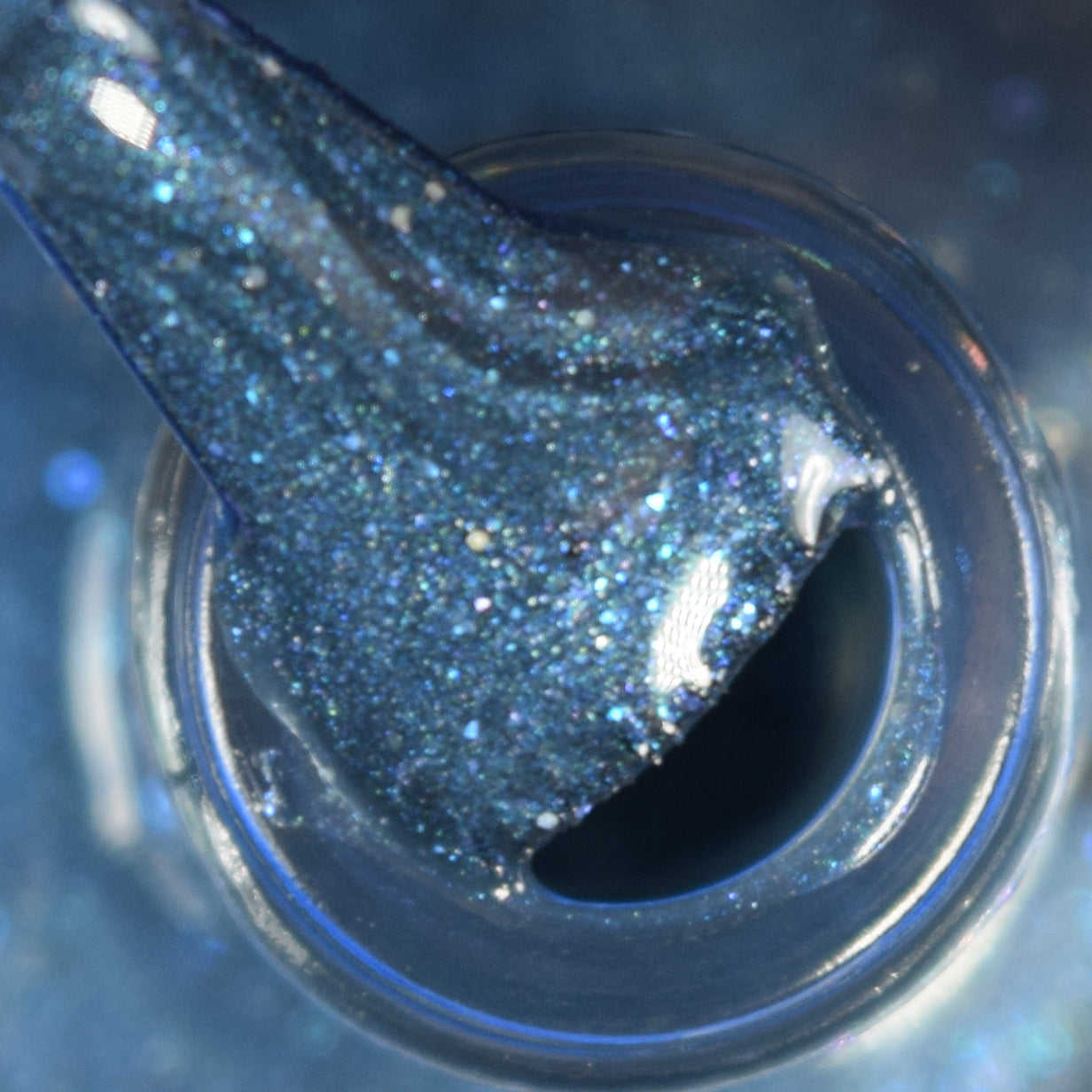 A close-up of a PI Colors Tanager.043 Blue Nail Polish bottle with a brush coated in shimmering teal glitter polish. The bristles are visibly wet and sparkling under bright light, creating a glossy effect. The background is softly blurred, enhancing the captivating allure of this stunning hue.