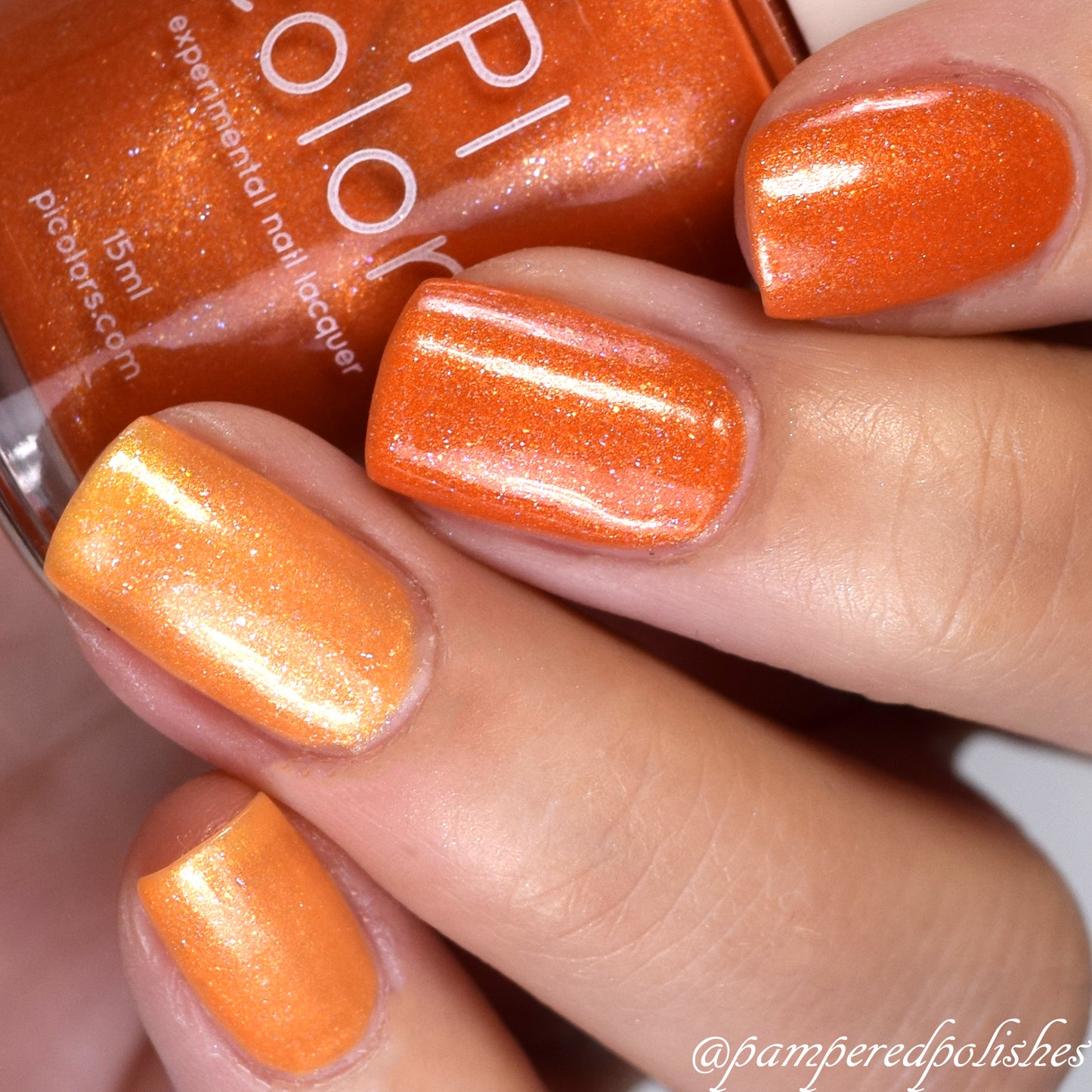 A hand with manicured nails showcases a shimmering luxury orange hue from the Sun Conure.097 Orange Nail Polish by PI Colors. In the background, the partially visible bottle highlights its 10-free formula on the label. The nails gleam with a glossy, metallic finish that beautifully catches the light.