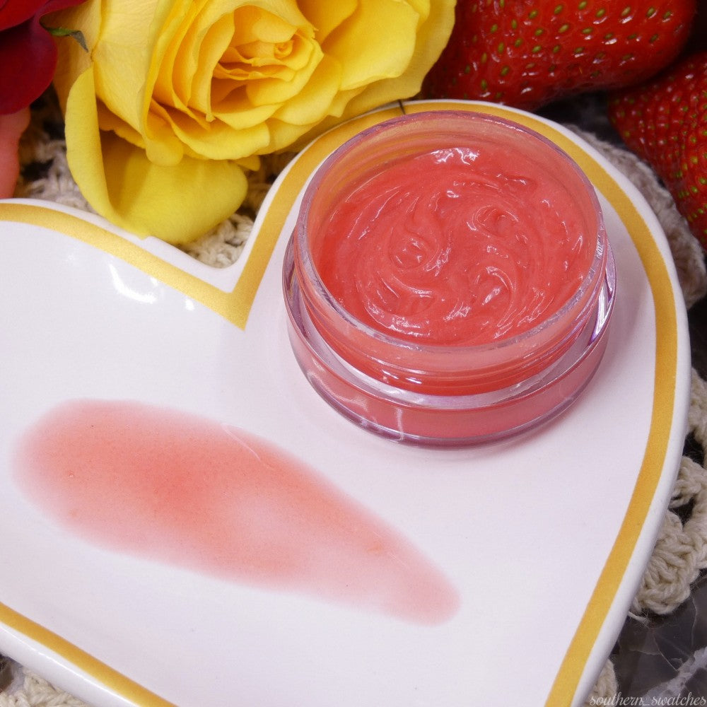 On a heart-shaped dish rests a small jar of PI Colors' Strawberry Scented Cuticle Butter, with a smear beside it. The vibrant yellow roses and strawberries in the background add a colorful touch to the setting. Infused with organic butters and healing oils, this cuticle butter nourishes while releasing delightful strawberry scents.