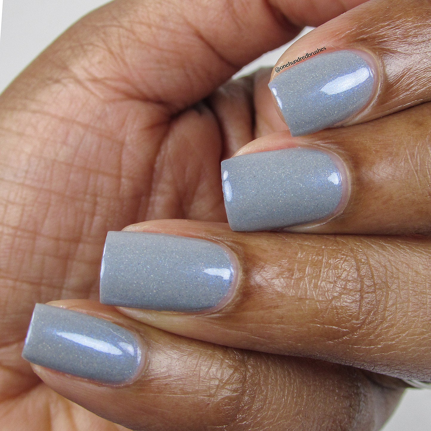 Close-up of a person's hand with short, square-shaped nails painted in Draco.055 Blue Gray Nail Polish by PI Colors, boasting a glossy light gray shade with subtle holographic glitter. The skin tone is a rich brown, and the hand is gently curled inwards.