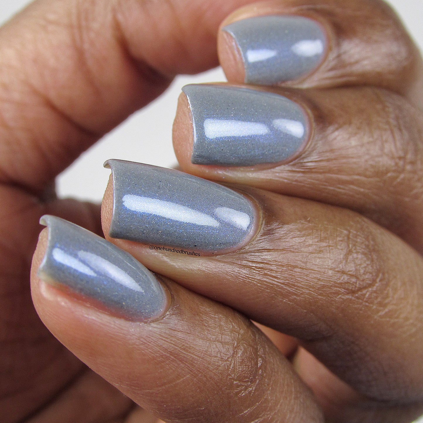A close-up of a person's hand with short, neatly manicured nails showcases the dazzling Draco.055 Blue Gray Nail Polish by PI Colors. The shimmery light blue with holographic glitter elegantly complements the rich brown skin tone, enhancing its glossy, sparkling finish.