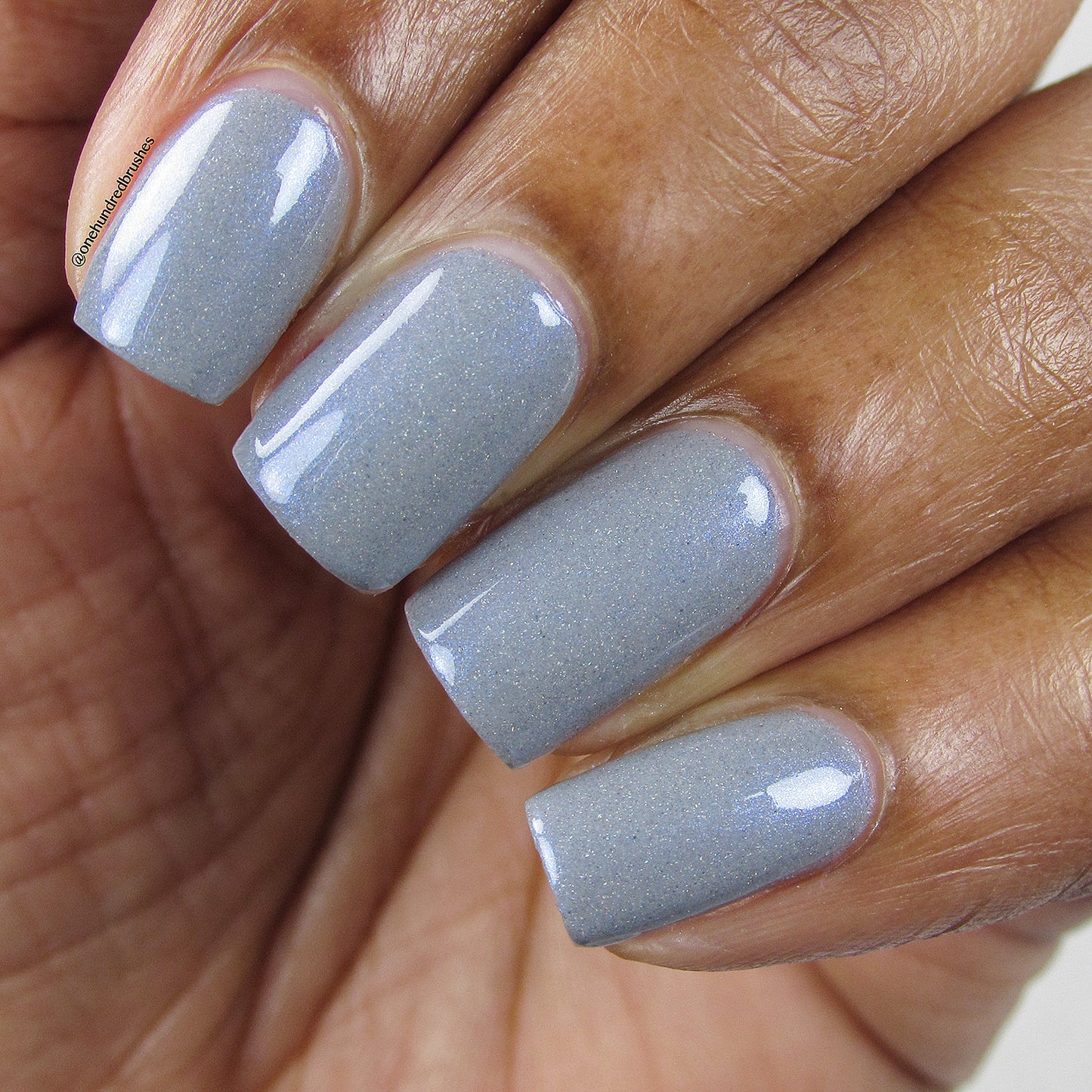 A close-up of a hand with neatly manicured nails painted in Draco.055 Blue Gray Nail Polish by PI Colors, displaying a glossy, light gray color enhanced with subtle holographic glitter. The skin tone is medium brown, and the nails have a square shape with a smooth finish.