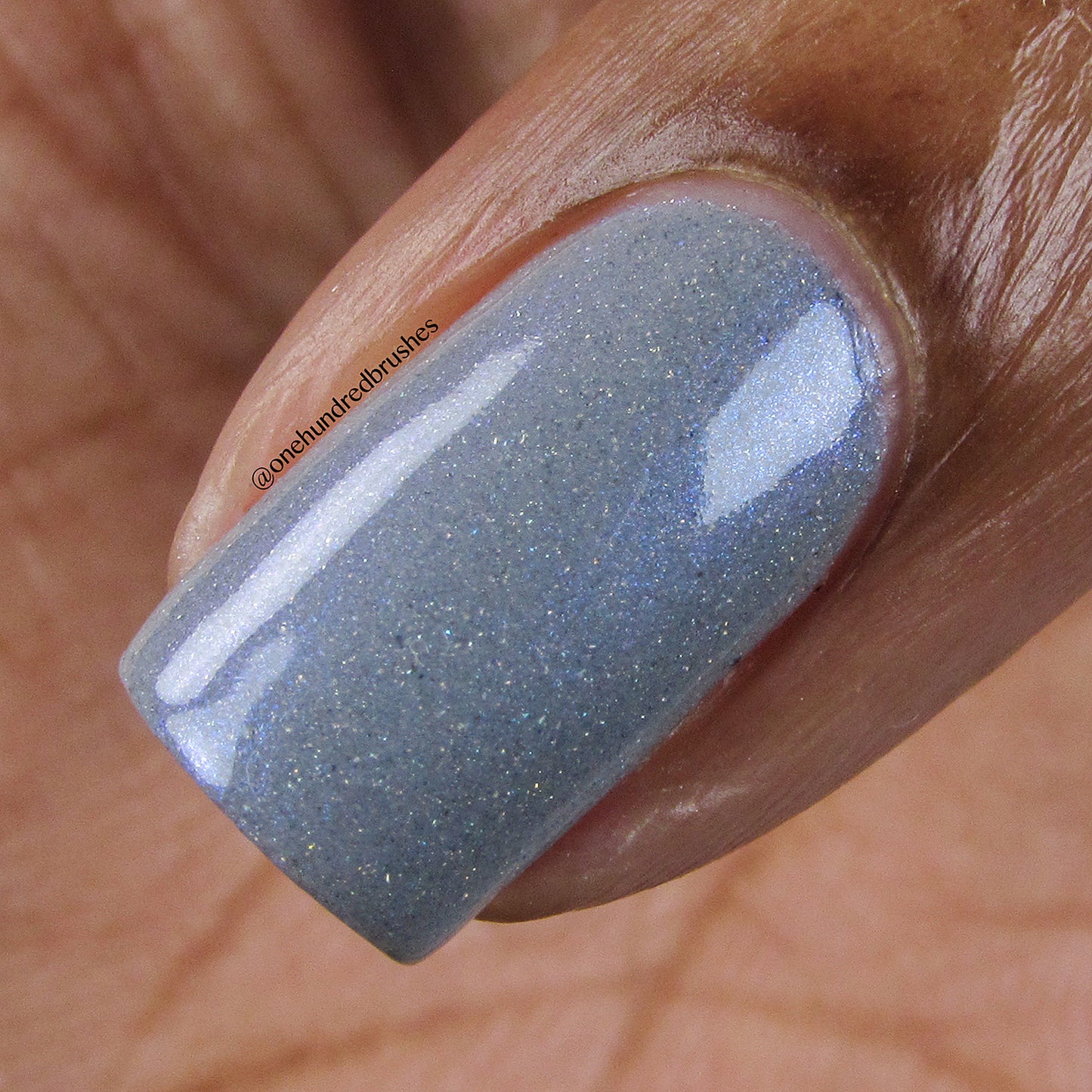 A close-up of a hand displays a single fingernail adorned with PI Colors' Draco.055 Blue Gray Nail Polish, boasting a glossy, shimmery greyish-blue hue. The nail's surface captures light beautifully, highlighting the polish's holographic glitter and smooth texture.