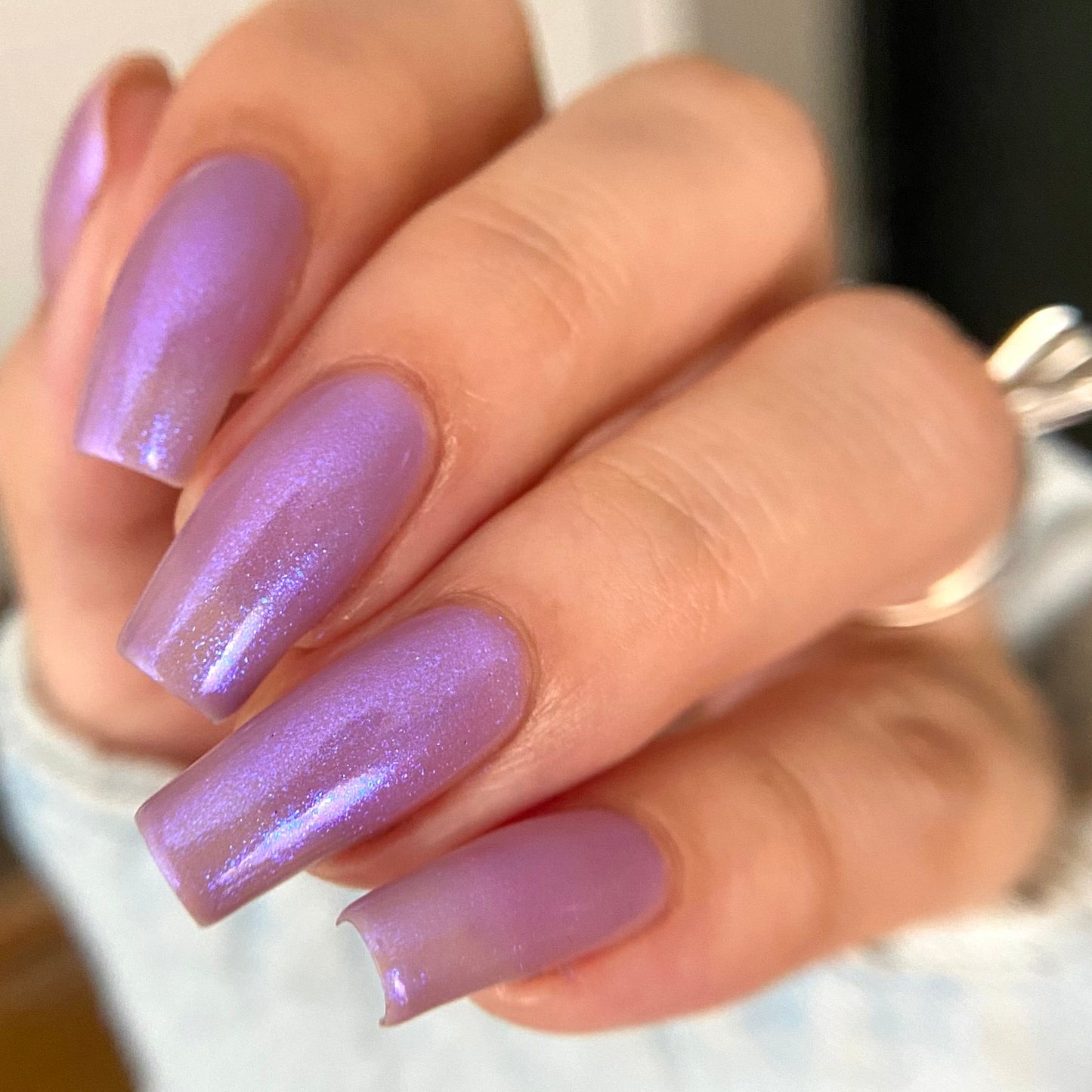 A close-up of a hand with long, almond-shaped nails painted in a shimmering light purple/lavender polish, wearing a silver ring.