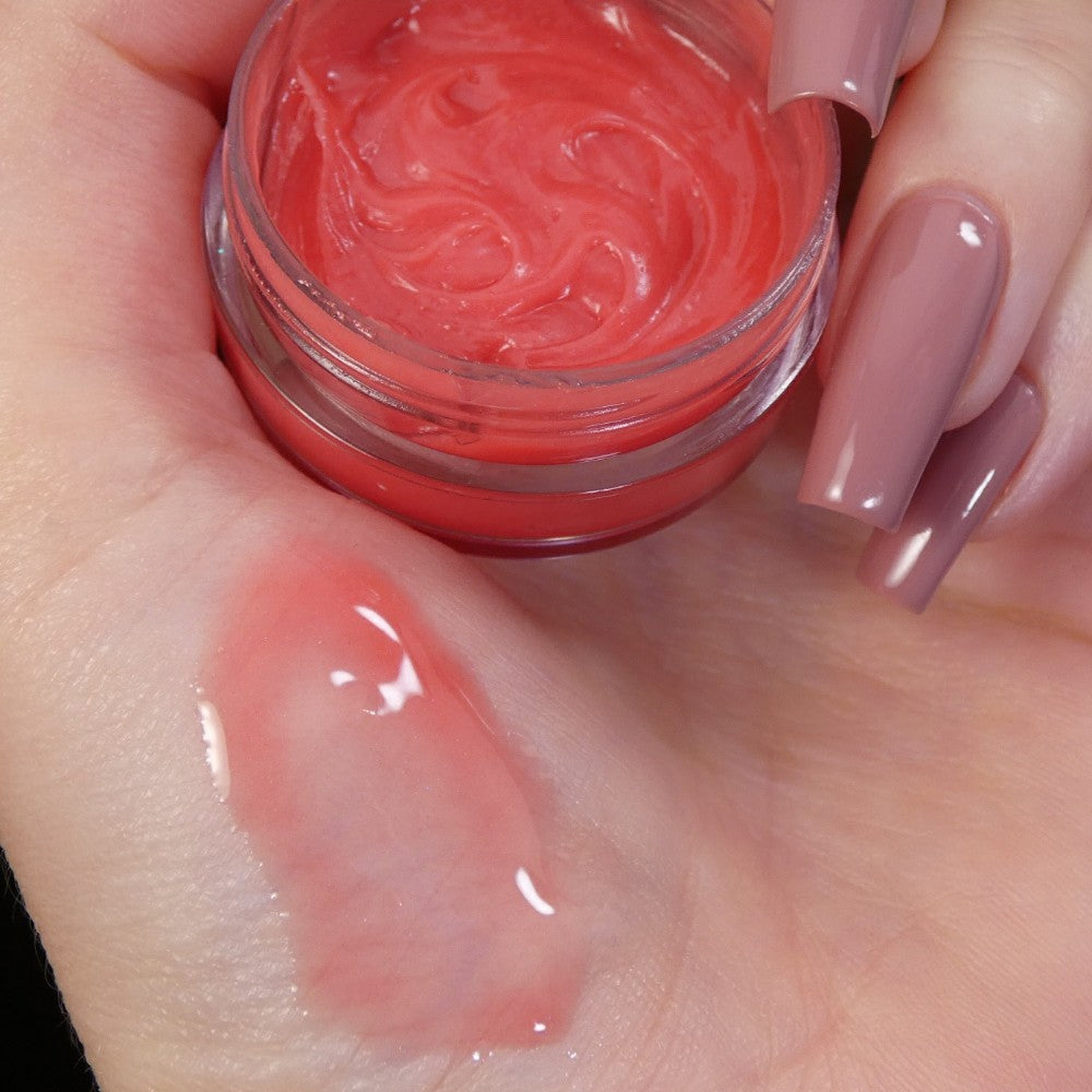 A hand with long, pinkish-beige painted nails holds an open jar of Strawberry Scented Cuticle Butter from PI Colors. Enhanced with natural oils, a dab of the butter is displayed on the palm, showcasing its glossy texture.
