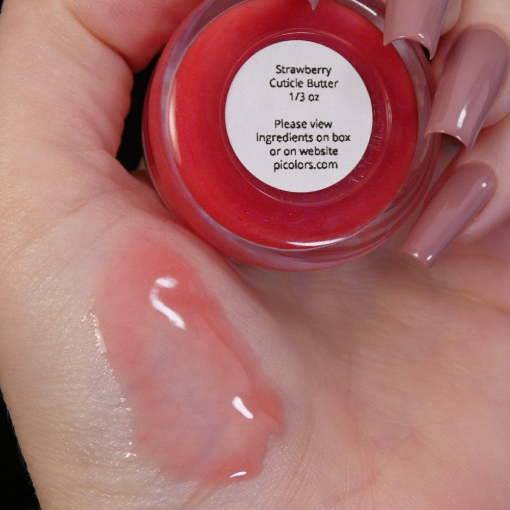 A neatly manicured hand holds a small open jar of PI Colors' "Strawberry Scented Cuticle Butter." The jar, filled with a luxurious blend enriched with natural oils, is labeled, and you can see some of the cuticle butter smeared on the back of the hand.