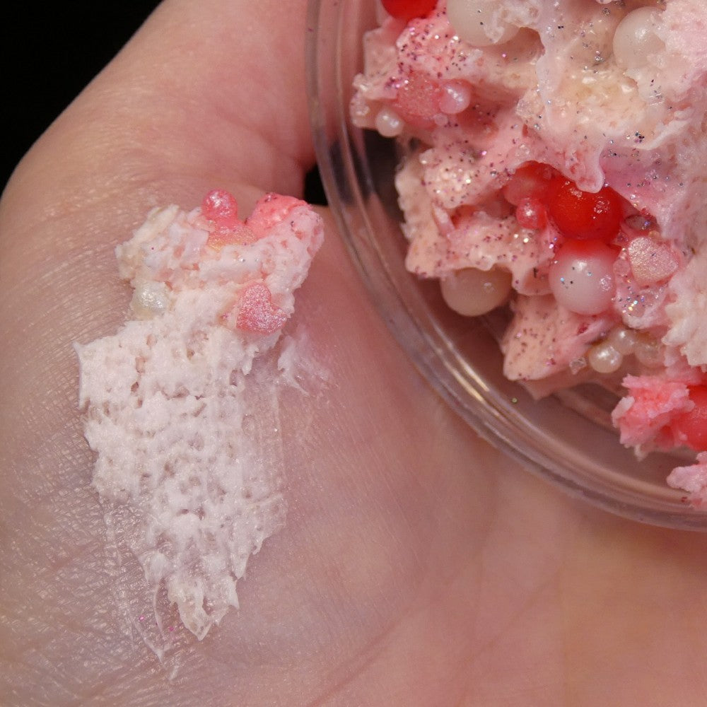 A hand holding a bowl containing Strawberry Whipped Soap Sugar Scrub by PI Colors, showcasing its textured, colorful appearance akin to strawberry whipped soap. The scrub, with hues of pink and red interspersed with small, round beads and sparkling glitter, spreads onto the fingers, highlighting its sticky consistency.