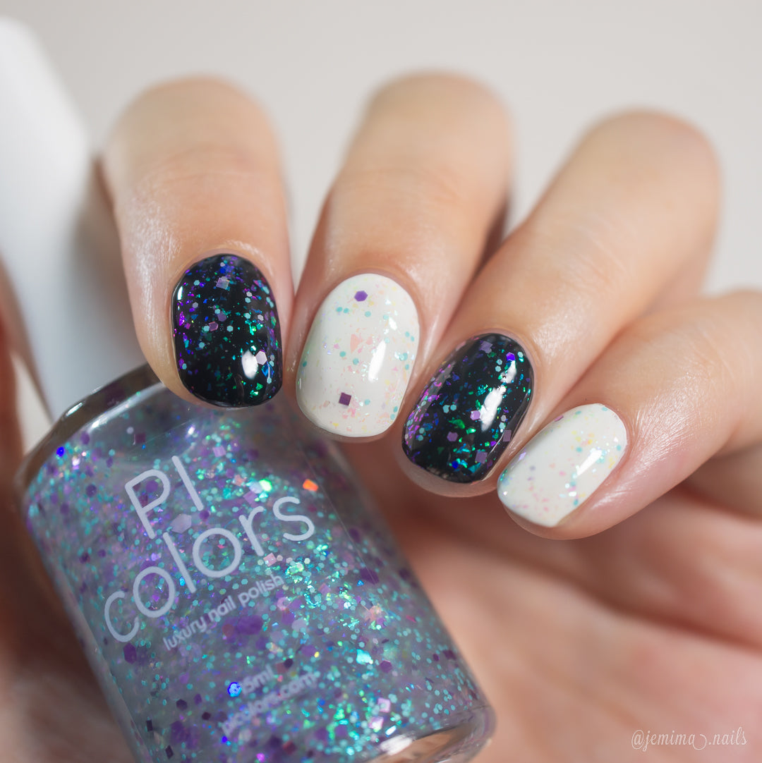 A hand displays vibrant nail art: two nails sparkle with blue and green glitter, while three showcase a white base adorned with multicolored speckles. In the background, a bottle of PI Colors' Hyacinth Rains.000 nail polish is visible, ideal for use as a topper with its colorshifting rainbow flakies and 10-free formula.