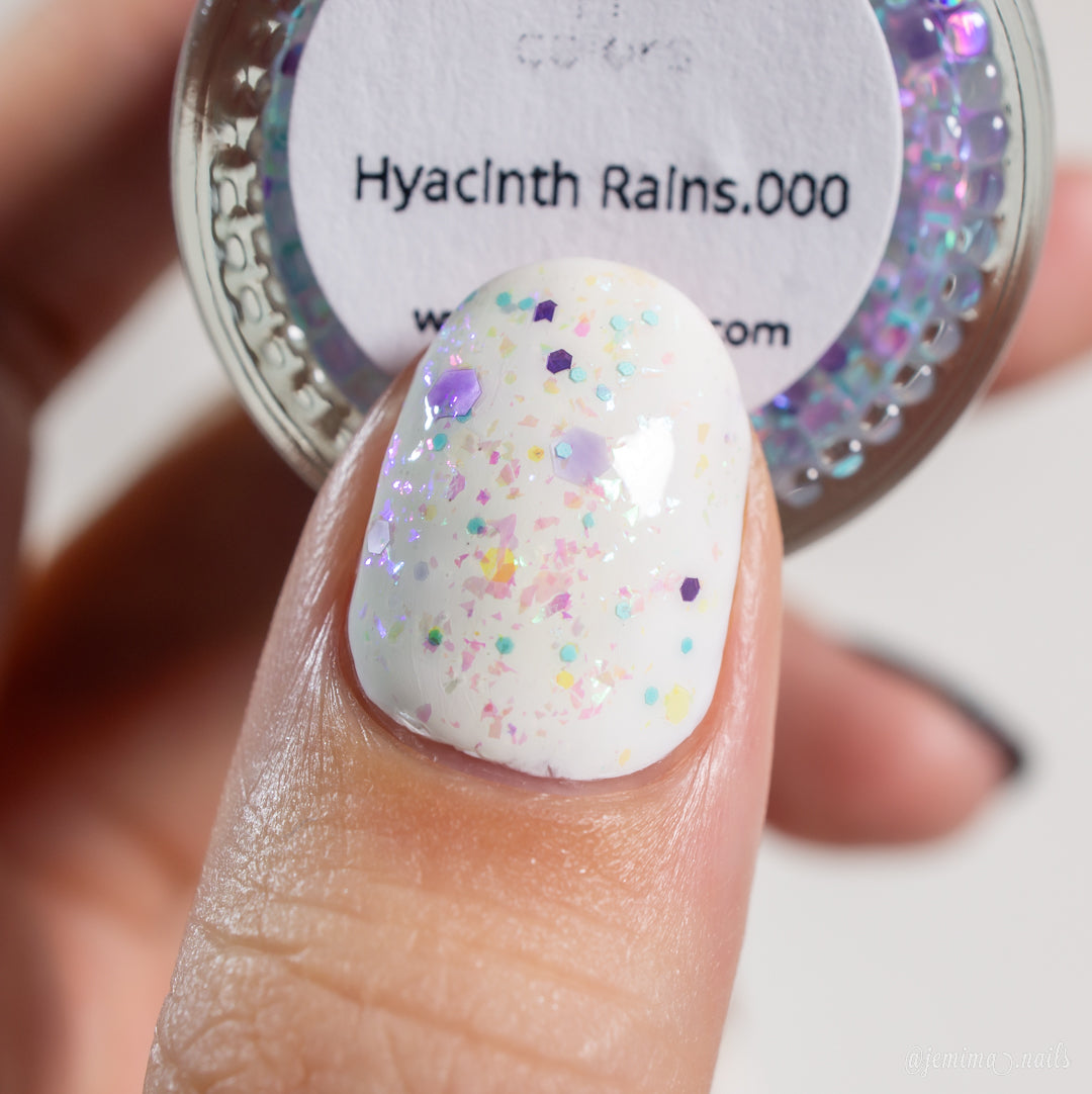 A close-up of a finger adorned with white nail polish showcasing vibrant confetti-like specks. In the background, a small container labeled "Hyacinth Rains.000" by PI Colors can be seen, containing similar sparkling particles and 10-free color-shifting rainbow flakies for an enchanting nail polish topper effect.