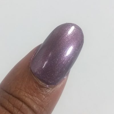 A close-up image of a fingertip adorned with PI Colors' Cœur.005 Purple Nail Polish showcases its shimmery plum hue. A plain, light-colored background emphasizes the vivid elegance of this striking shade.