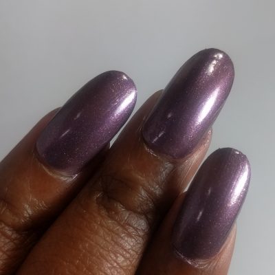 Close-up of a hand with dark skin displaying nails painted in PI Colors Cœur.005 Purple Nail Polish, a glossy and metallic plum shade. The background features a light, blurred surface. The nails are rounded and smooth, shimmering subtly as if adorned with glitter.