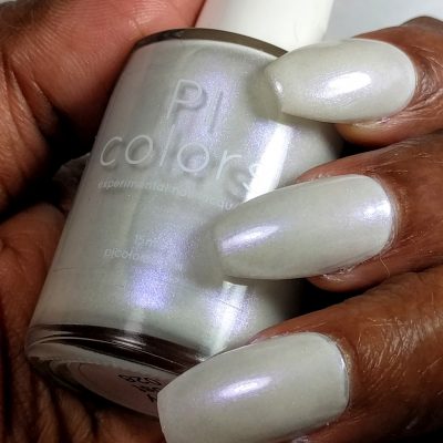 A person holds a bottle of PI Colors' Candy Frost.026 White Nail Polish, renowned for its pale iridescent sheen enhanced with holographic powder. Their fingernails are painted to match the bottle's shimmering light hue, showcasing the enchanting sparkle of this 10-free formula.