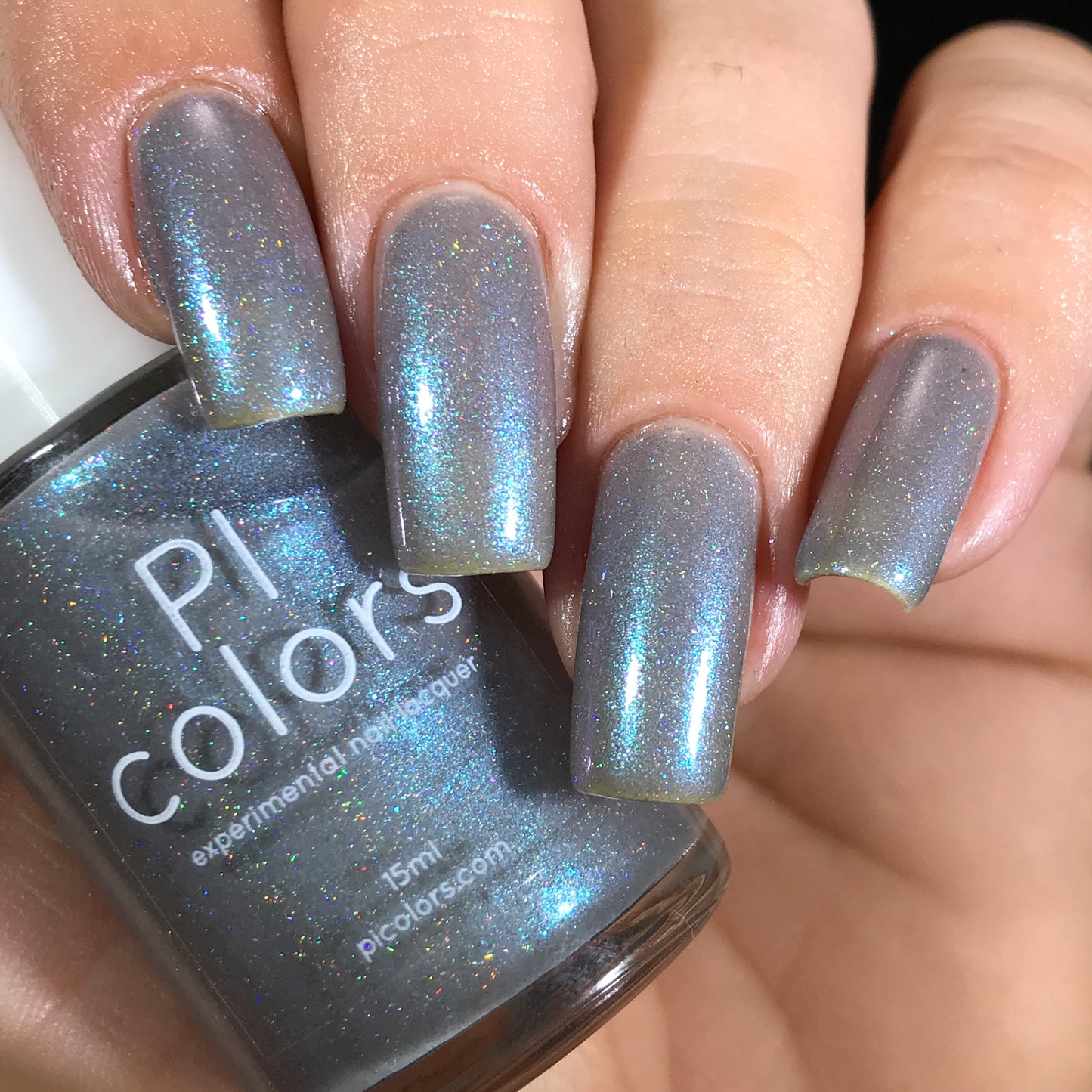 A hand displays long nails adorned with a shimmery, holographic blue gray polish. The bottle of "Asperitas.105 Blue Gray Nail Polish" from PI Colors is held between the fingers, its holographic sparkle reflecting various colors in the light.