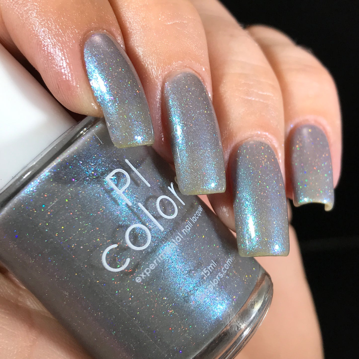 A hand with long nails painted in a shimmery metallic blue-gray nail polish is holding a bottle of the same handmade polish. The bottle is labeled "PI Colors," and the glittery finish reflects light, offering a holographic sparkle that enhances its allure.