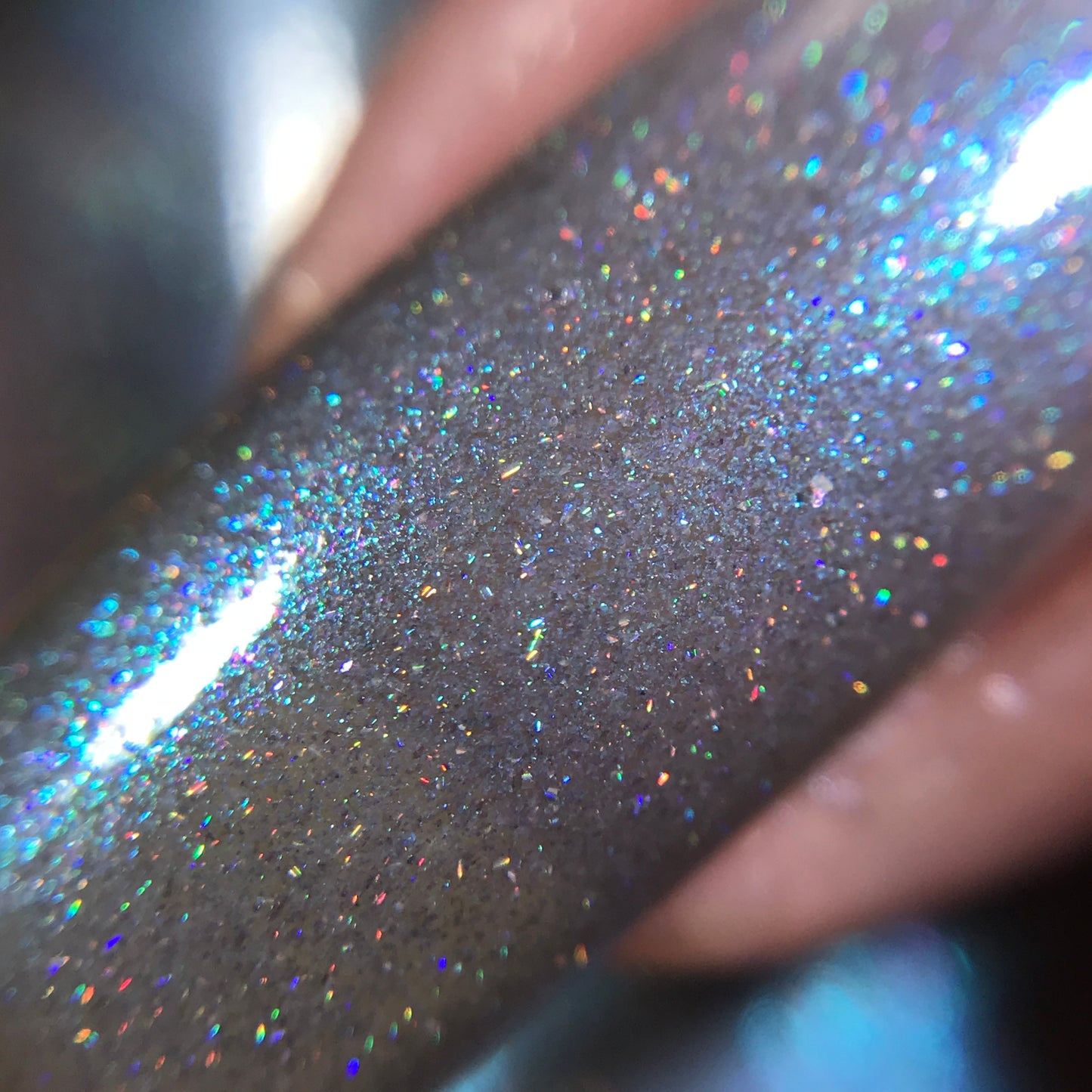 Close-up of a nail featuring PI Colors' Asperitas.105 Blue Gray Nail Polish with handmade holographic glitter. The surface shimmers with a blend of silver and iridescent colors, reflecting an array of hues such as blue, green, and pink when illuminated.