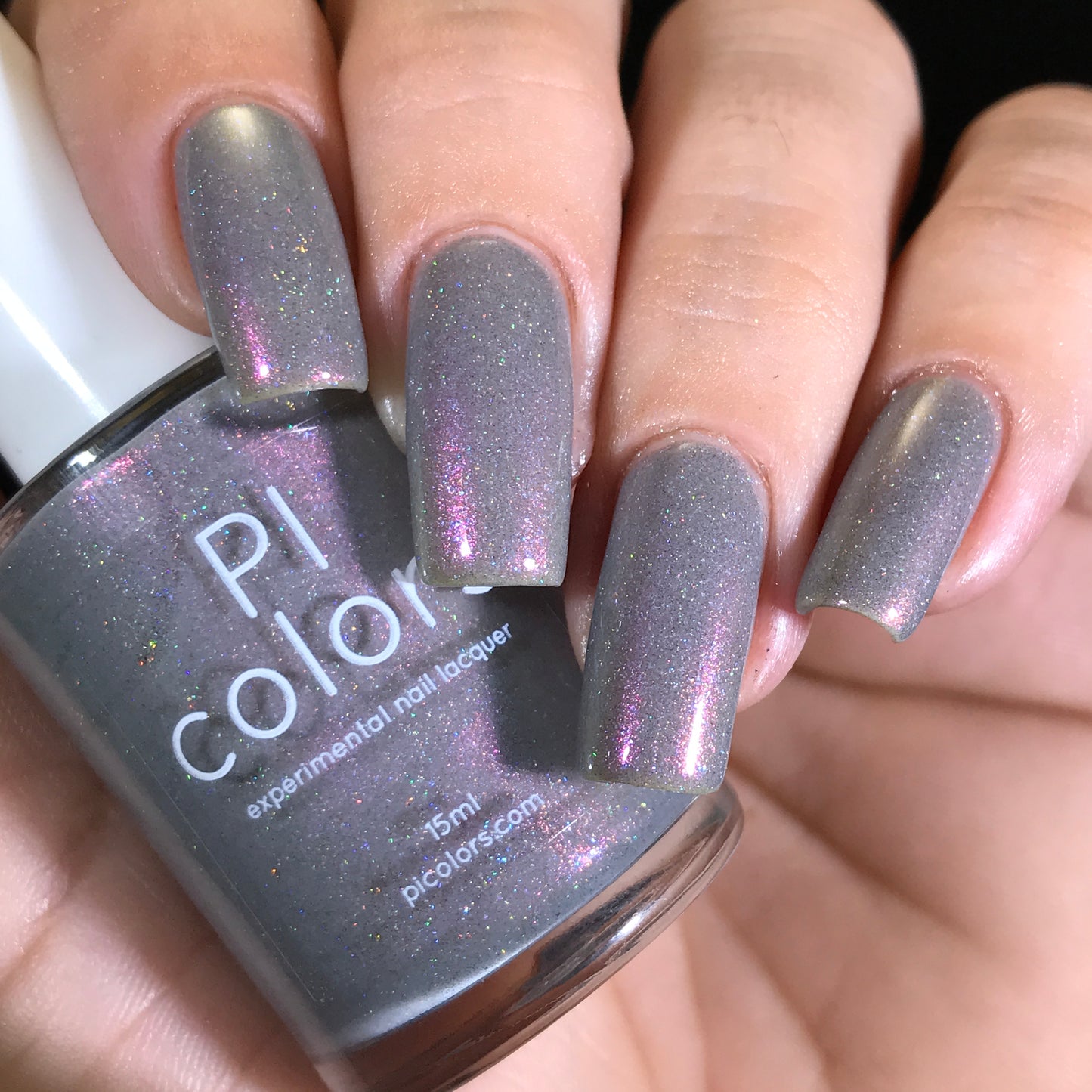 A hand with long nails painted in Cirrus.118, a gray-based polish featuring 10-free, pink, and purple shimmer hues, holds a bottle labeled "PI Colors." The nail polish bottle perfectly matches the color on the nails.