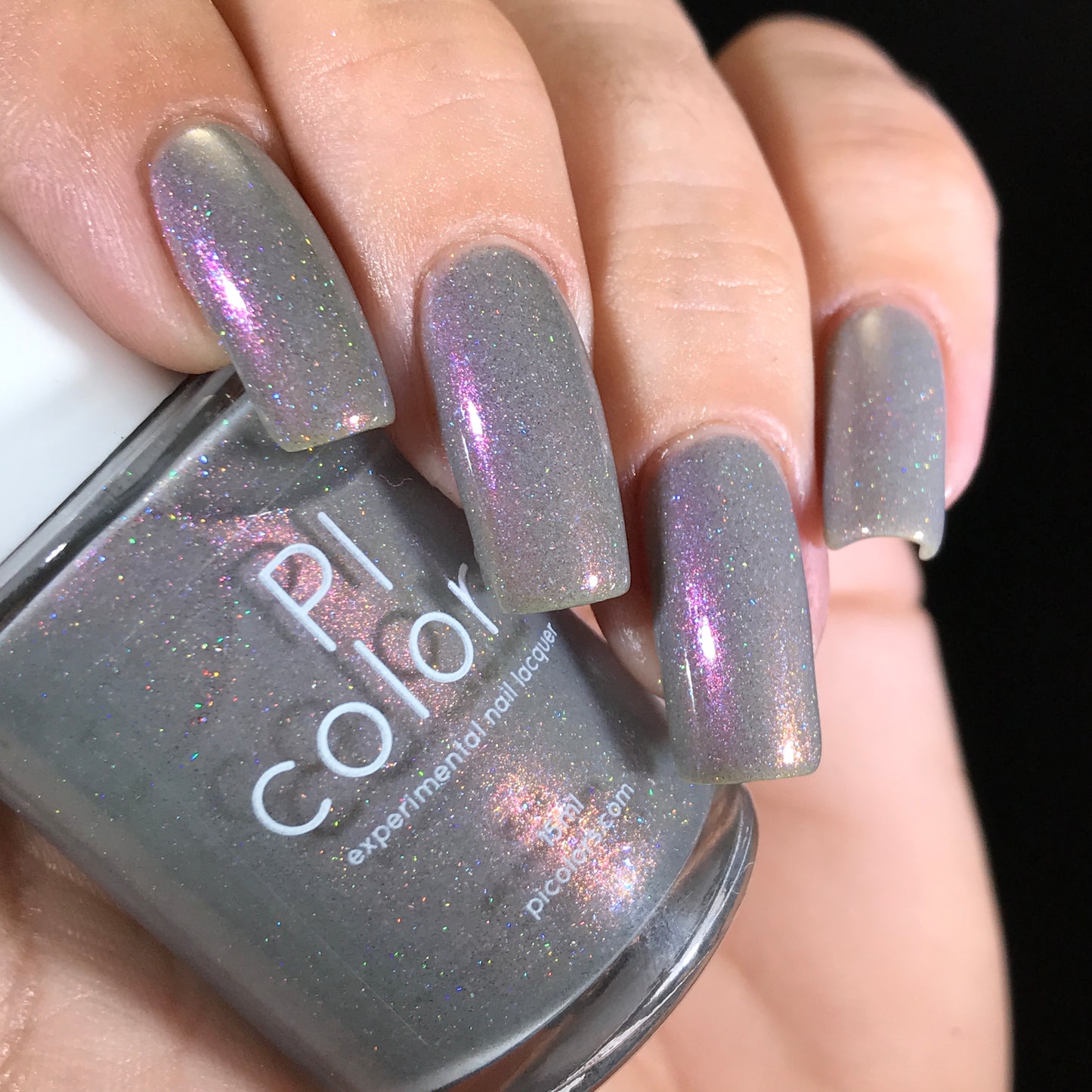 A hand with neatly manicured nails is displayed, painted in the Cirrus.118 Pink Gray Nail Polish, showcasing a subtle purple shimmer that sparkles as it catches the light. The hand clasps a bottle of 10-free nail polish labeled "PI Colors," perfectly matching the stunning manicure.