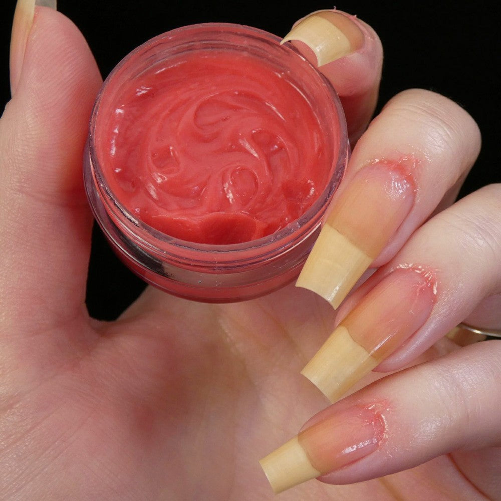 A hand with long natural nails holds an open container of PI Colors' Strawberry Scented Cuticle Butter. Some of the cream, enriched with natural oils, is on the fingertips surrounding the nails, indicating use for a manicure or therapeutic treatment. The background is black.