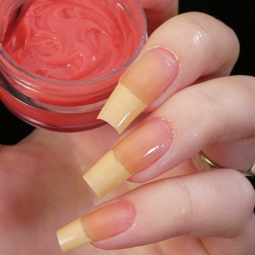 A close-up captures a hand with long, natural-toned nails holding a small open jar of PI Colors' Strawberry Scented Cuticle Butter, enriched with natural oils. The square-tipped nails appear polished or coated with a clear layer, set against a dark background that highlights their elegant simplicity.