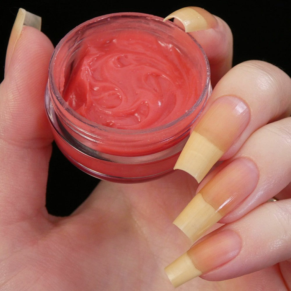 A hand with long, natural nails holds a small open container of red gel. The nails have a clean, glossy appearance as they hover over PI Colors' Strawberry Scented Cuticle Butter. The background is black, accentuating the vibrant color and creamy texture of this indulgent product.