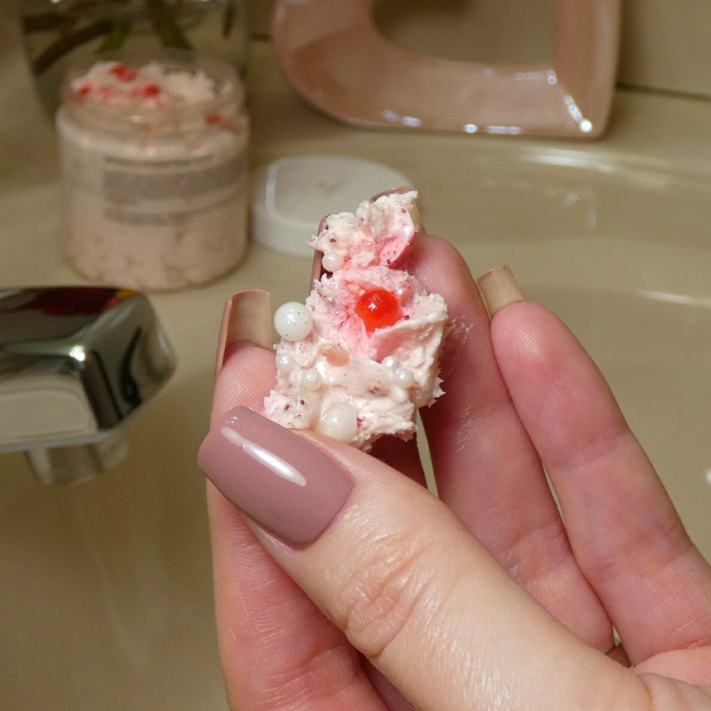 A hand with pink painted nails holds a small amount of PI Colors' Strawberry Whipped Soap Sugar Scrub, featuring red beads and white pearls. A jar of this creamy pink substance rests on the sink, enriched with the nourishing essence of avocado oil.