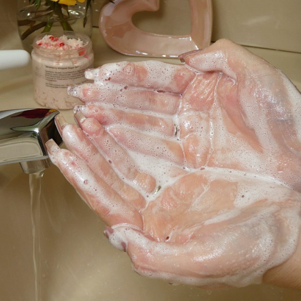 Hands cupped under a faucet, lathered with the Strawberry Whipped Soap Sugar Scrub by PI Colors, with water running. A jar of this soap sugar scrub is visible in the background, alongside a heart-shaped decoration on a countertop.