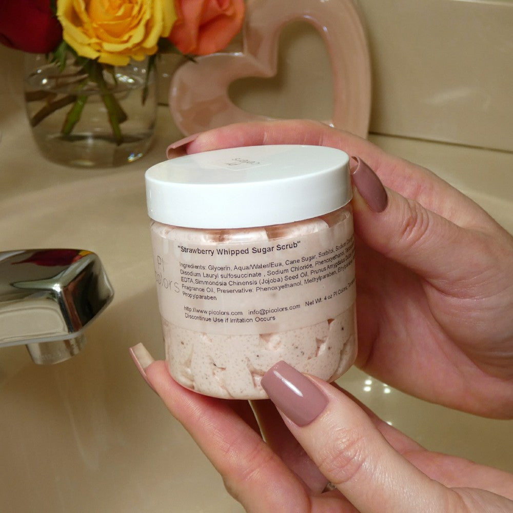 A person with manicured nails holds a jar of "Strawberry Whipped Soap Sugar Scrub" by PI Colors near a bathroom sink. The scrub is pale pink, and vibrant flowers bloom in the background, enhancing the serene atmosphere.