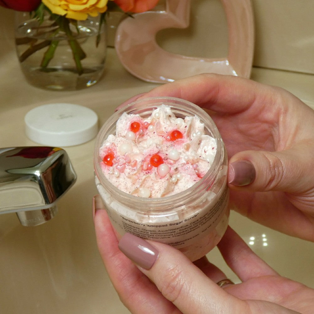 A person holding an open jar of PI Colors' Strawberry Whipped Soap Sugar Scrub, adorned with red and pearlescent beads, stands next to a faucet. In the background, there's a vase with flowers and a decorative heart.