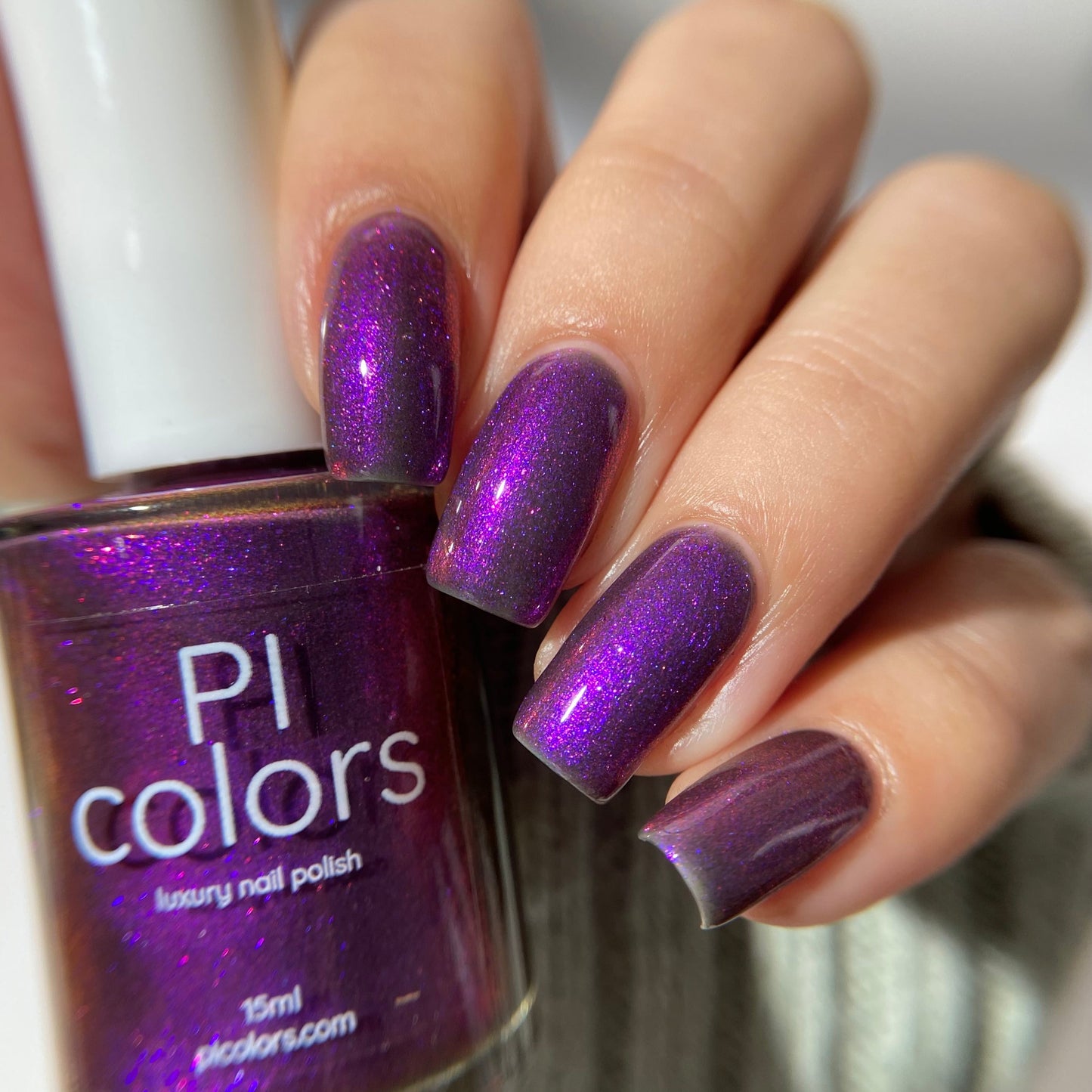 A hand with nails painted in shimmering purple multichrome polish holds a bottle labeled "Danbury Does It.000" by PI Colors. The 10-free, vibrant, glossy nails and bottle are set against a blurred neutral background.