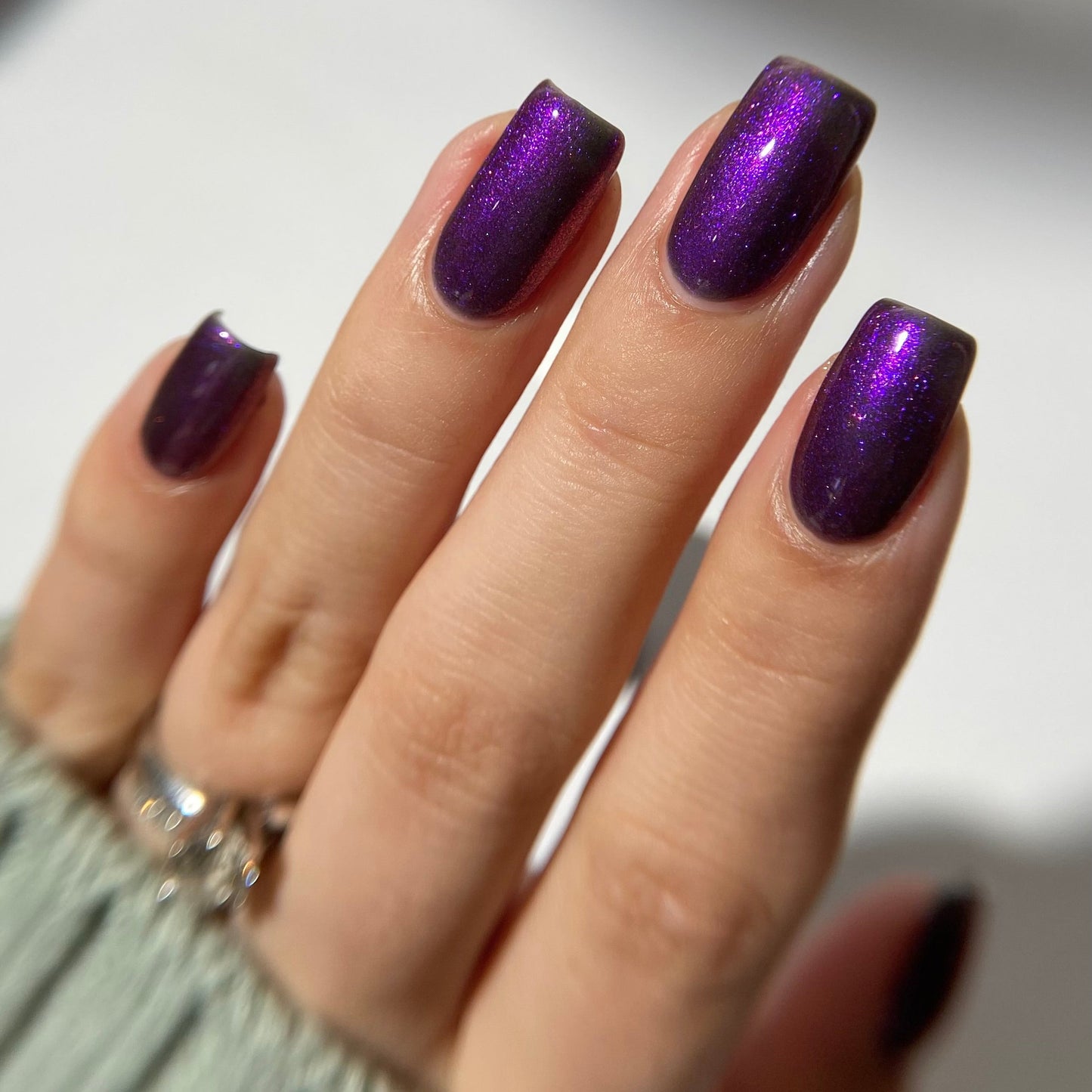 A hand showcases medium-length, square-shaped nails painted with PI Colors' Danbury Does It.000, a glossy purple multichrome polish. The silver ring and light gray ribbed sweater enhance the elegant look against a blurred white backdrop that highlights the nail lacquer's 10-free finish.