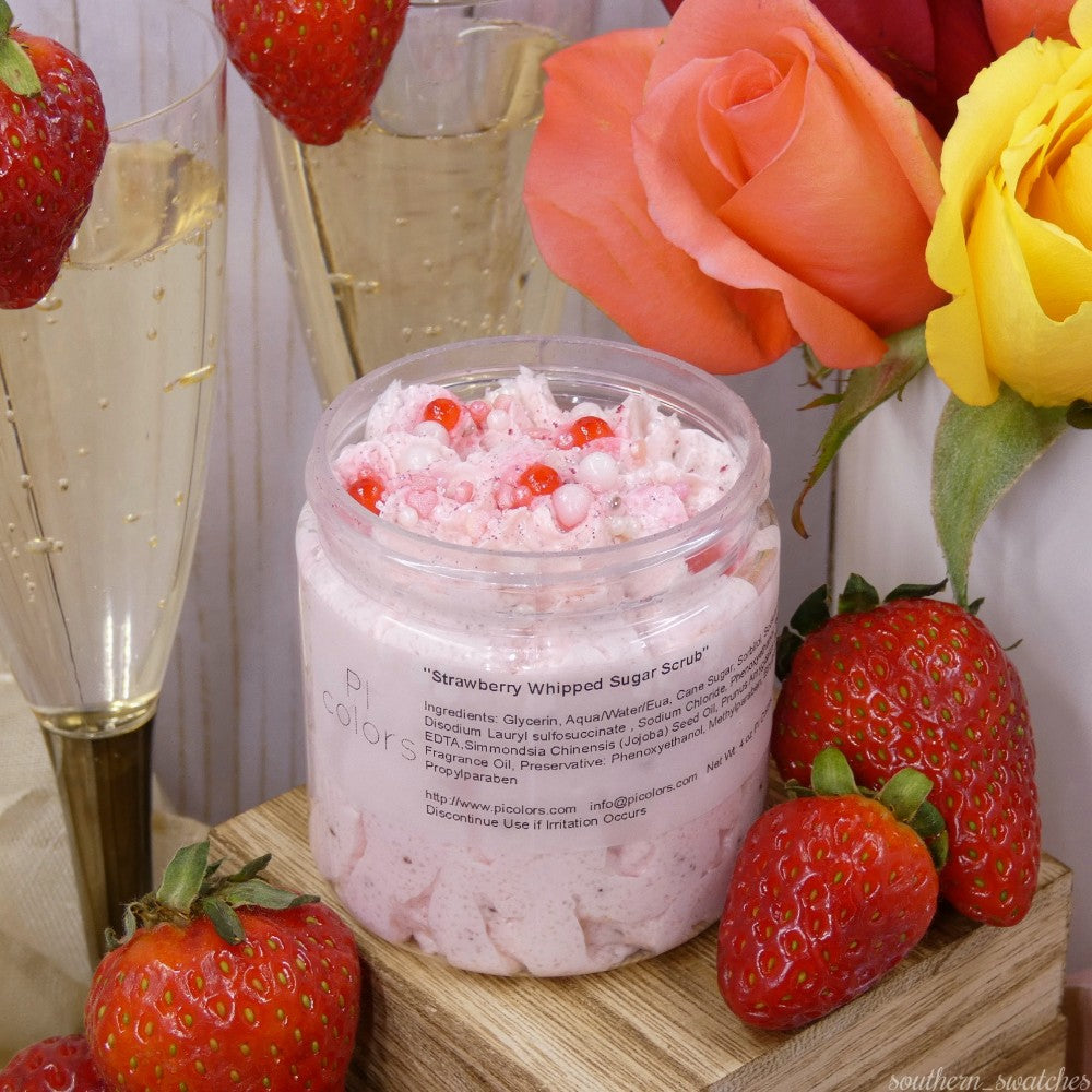 A jar of PI Colors' Strawberry Whipped Soap Sugar Scrub is surrounded by fresh strawberries and two champagne glasses, with a touch of avocado oil in the background. The scrub appears pink with small red beads, and the label displays its ingredients. A yellow cloth slightly enters the frame from the right.