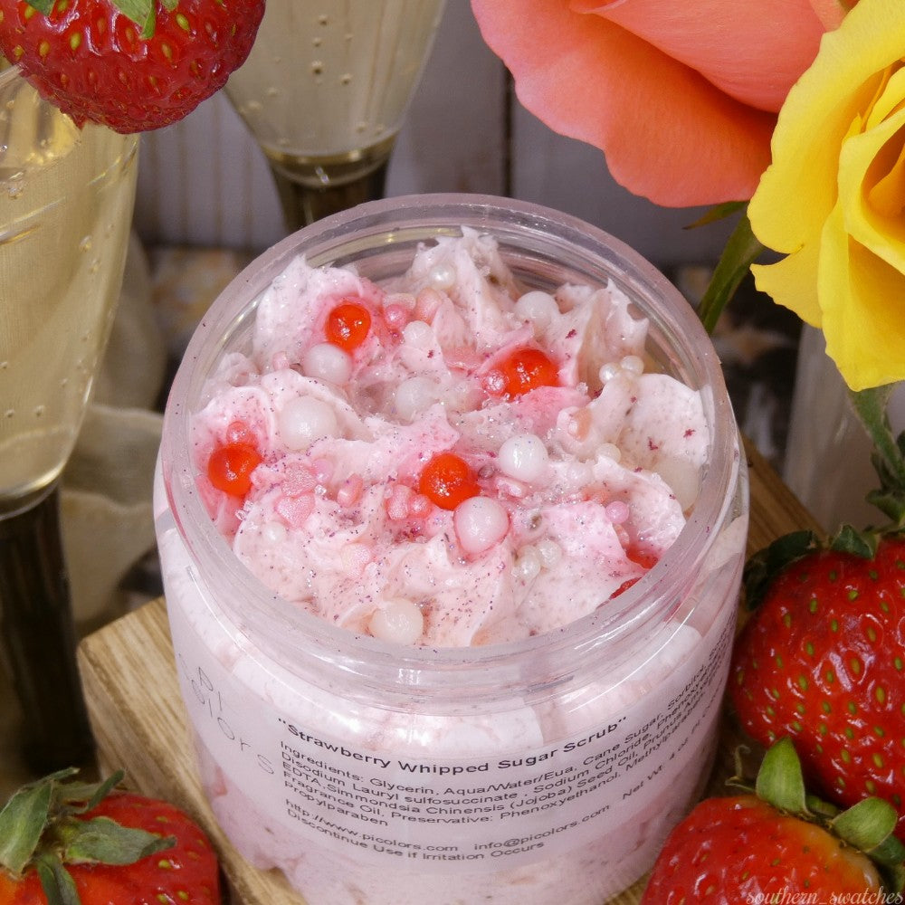 An open jar of PI Colors' Strawberry Whipped Soap Sugar Scrub, embellished with red and white beads, rests amidst fresh strawberries and roses. Two champagne glasses in the background lend an air of elegance, enhancing the luxurious experience enriched with nourishing avocado oil.