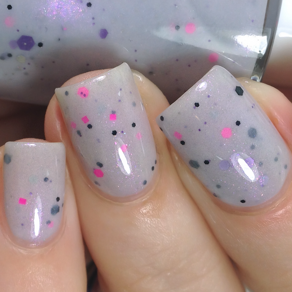 Close-up of nails painted with PI Colors' Kirin.000 Lilac Purple Nail Polish, showcasing a pale grayish purple crelly adorned with small black, pink, and purple speckles. The glossy finish enhances the elegance, while the partially visible bottle in the background suggests the quality of this handmade nail polish.