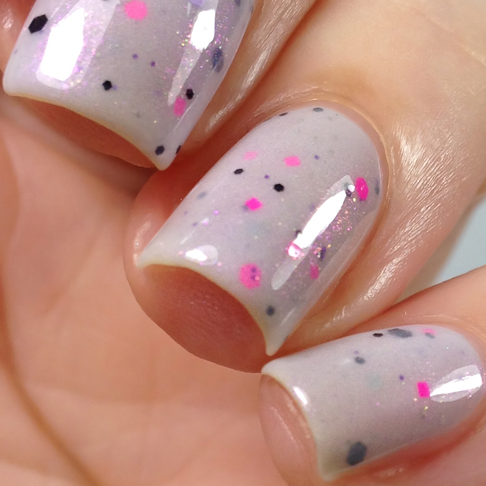A close-up reveals fingernails adorned with PI Colors' Kirin.000 Lilac Purple Nail Polish, a crelly with pink, black, and purple glitter flecks. The nails boast a smooth and glossy finish that emphasizes the distinct shimmer against a softly blurred backdrop.