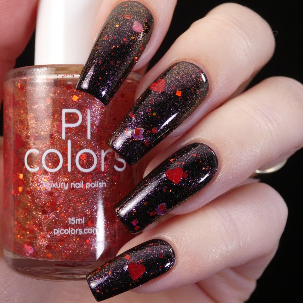 A close-up shows a hand showcasing long, nails adorned with black glittery nail polish featuring red, holographic hearts. The hand holds a bottle of PI Colors' Strawberry Heart.208 Red Nail Polish Topper, which is vibrant red with shimmering glitter and heart shapes embedded.