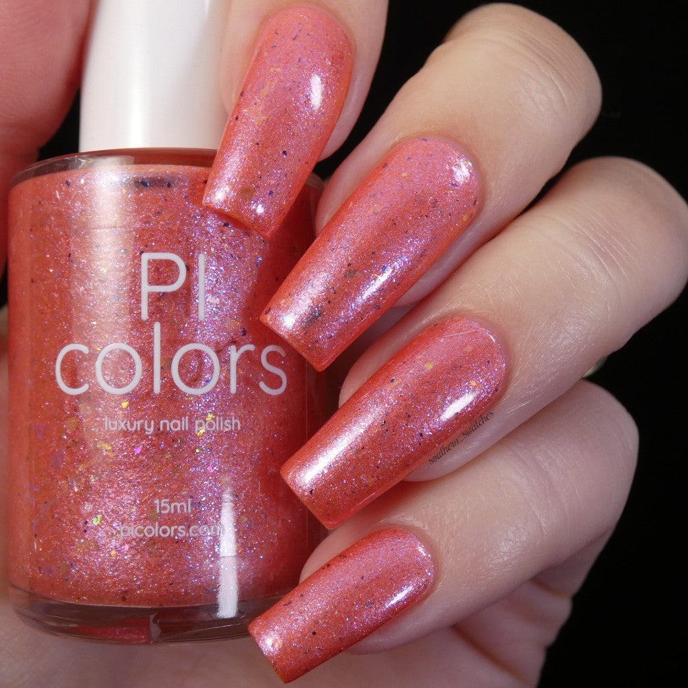 A hand with long nails is painted in a shimmery coral nail polish while holding a bottle of PI Colors' Lala Ngoxolo Kumkani Coral Nail Polish. The vibrant hue showcases iridescent rainbow flakes for a sparkly, dazzling finish.