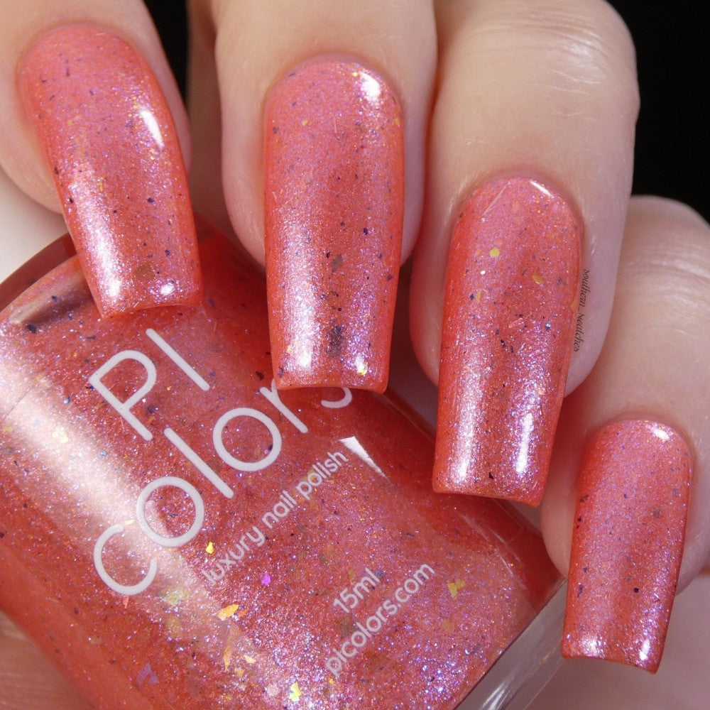 Close-up of hands displaying nails painted with a shimmering peachy coral nail polish. The "Lala Ngoxolo Kumkani Coral Nail Polish" by PI Colors comes in a matching bottle, revealing a shiny finish with reflective sparkles and iridescent rainbow flakes.