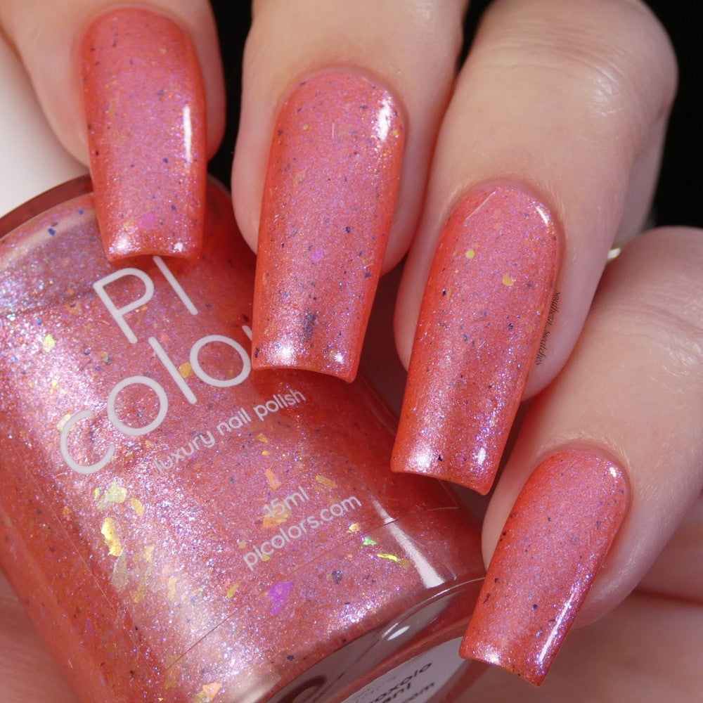 Close-up of hands with long nails painted in the luxury "Lala Ngoxolo Kumkani Coral Nail Polish" by PI Colors, a peachy coral color. The bottle of PI Colors is visible, highlighting an iridescent flakes nail lacquer that sparkles with a multicolored finish.